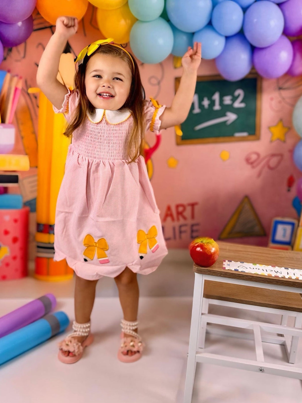 A young girl wearing the Girls Back To School Pencil Twirl Dress with bow appliques, smiling joyfully while standing in a colorful classroom setting with balloons, a chalkboard, and school supplies.
