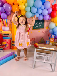 Young girl in a pink pencil plaid twirl dress with a bow and petal collar, posing amid colorful balloons.