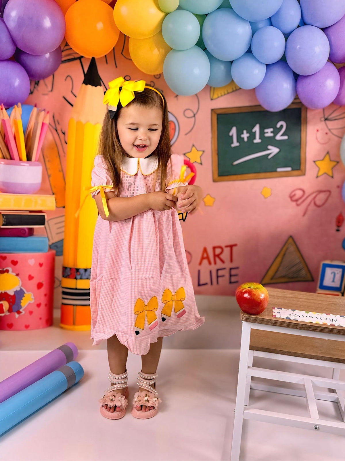 A cheerful girl in the Girls Back To School Pencil Twirl Dress, interacting playfully with school supplies in a festive classroom atmosphere filled with colorful balloons.
