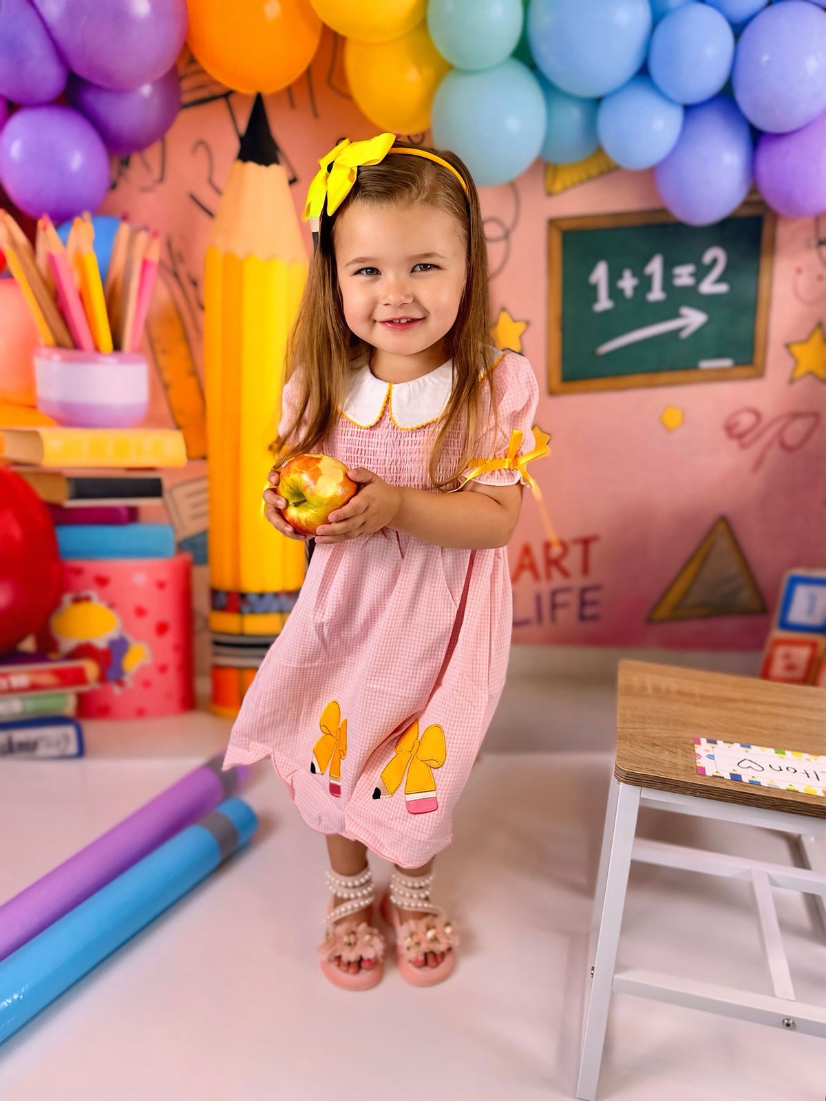 A young girl holding an apple, wearing the Girls Back To School Pencil Twirl Dress with delightful bow appliques, standing in a colorful classroom backdrop with balloons and educational elements.