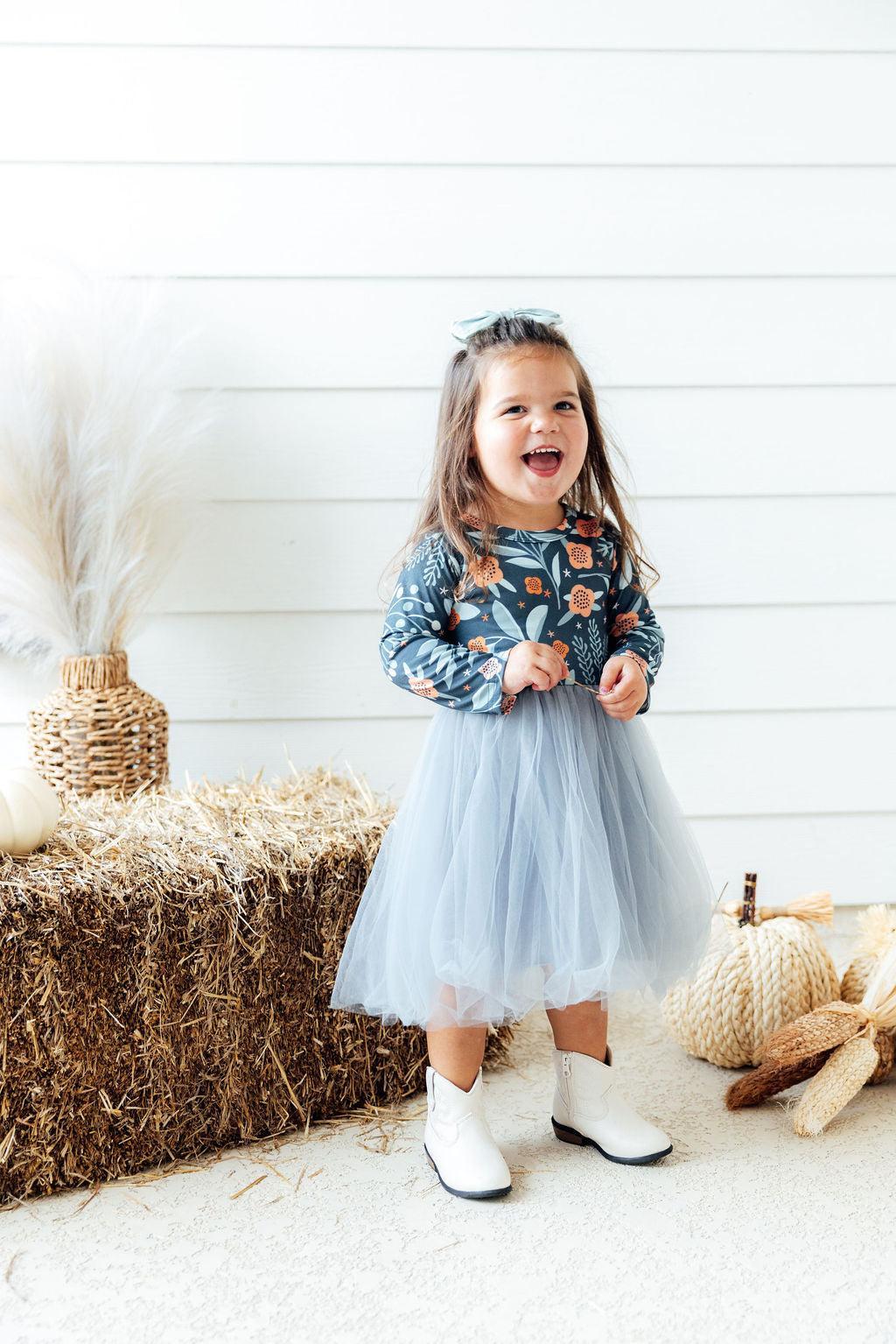 Toddler girl laughing in floral long sleeve bodice and light blue tulle tutu dress with white boots near hay bale.