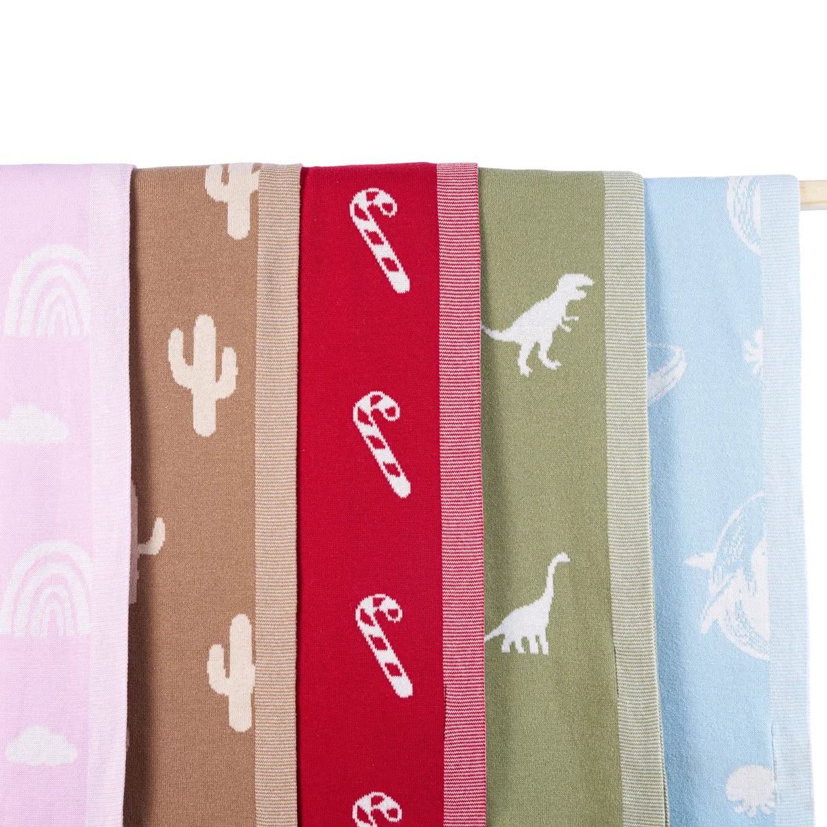 Five hanging baby blankets displaying lilac rainbows, brown cacti, red candy canes, olive dinosaurs, and blue marine life
