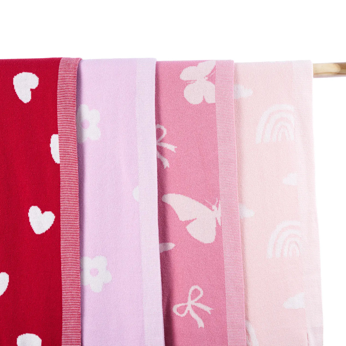 Four folded blankets hanging vertically on wooden rod, showing color gradient from red hearts to lavender daisies with ribbed