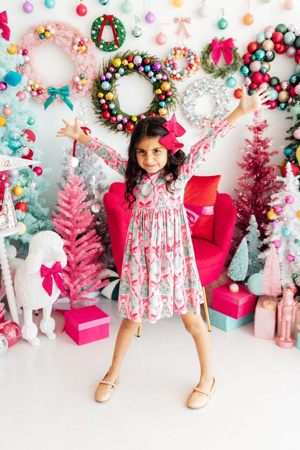 Girl posing with arms up in a pink long sleeve twirl dress with a holiday print, surrounded by colorful trees and wreaths.