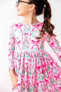 Girl in pink long sleeve dress with bow and wreath print, featuring a full circle skirt and front pocket for twirl-worthy fun.