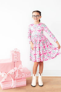 Girl in pink holiday print long sleeve twirl dress with pocket and white boots, standing beside stacked presents.