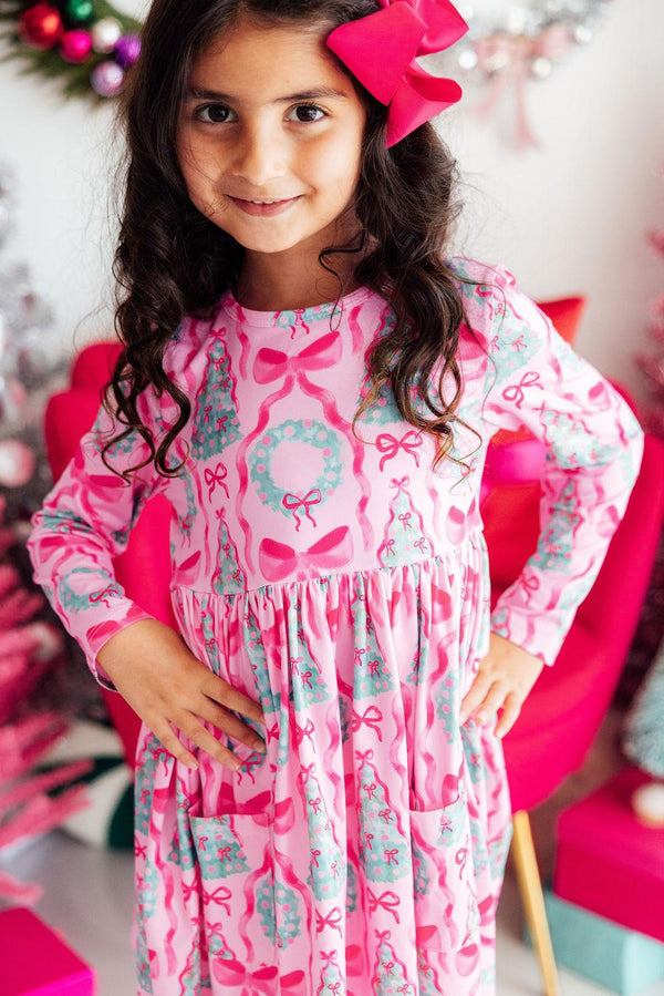Girl wearing a pink long sleeve twirl dress with bow and wreath print, front pockets, and a matching pink hair bow.