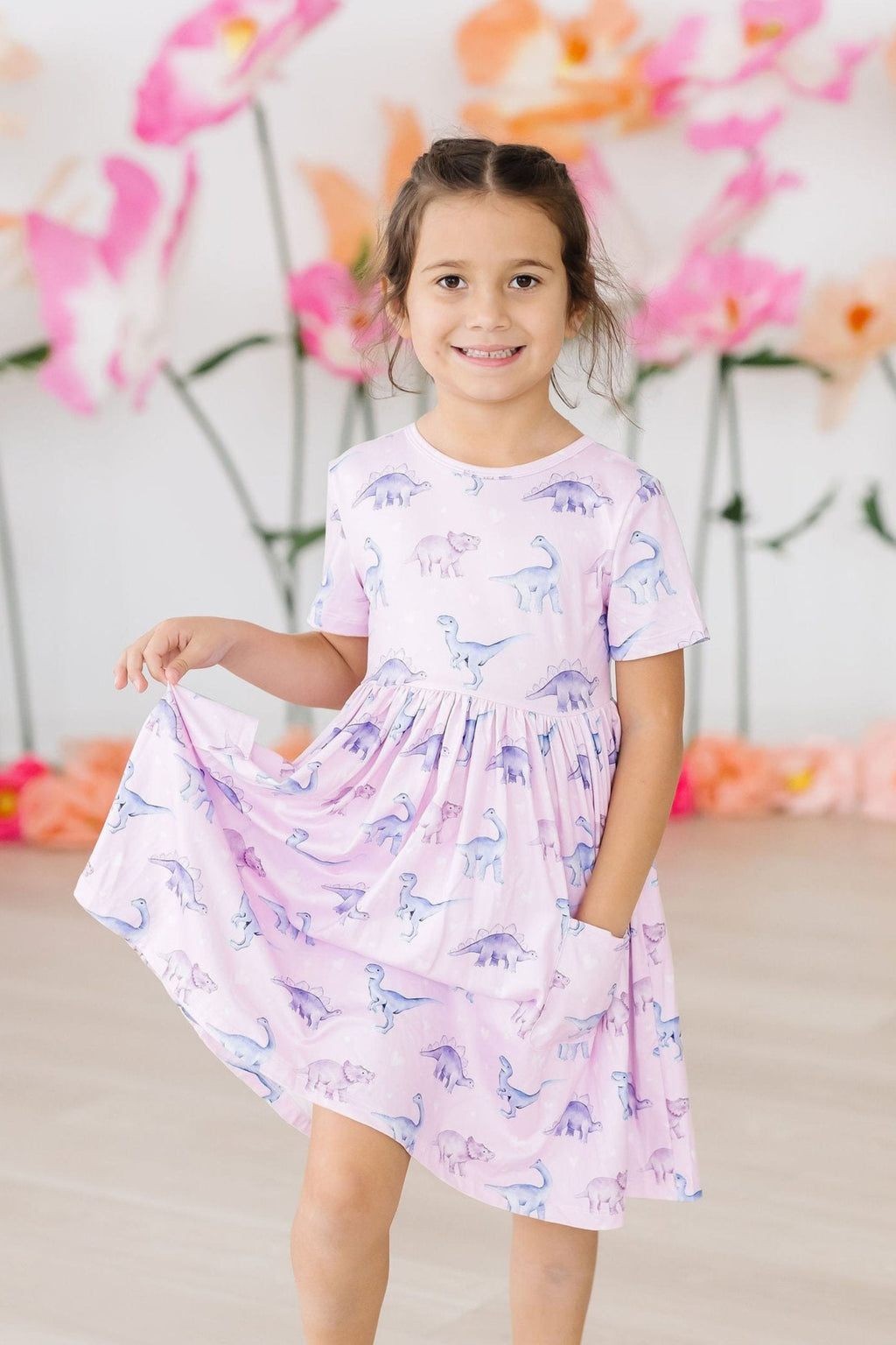Girl wearing the Jurassic Joy S/S Pocket Twirl Dress, showcasing a playful dinosaur print, twirling with joy in a vibrant and comfortable outfit perfect for kids' adventures.