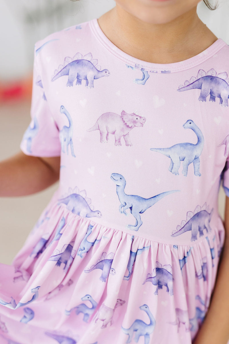 A child happily wearing the Jurassic Joy S/S Pocket Twirl Dress by Mila & Rose, showcasing a whimsical dinosaur print in soft pastel colors.