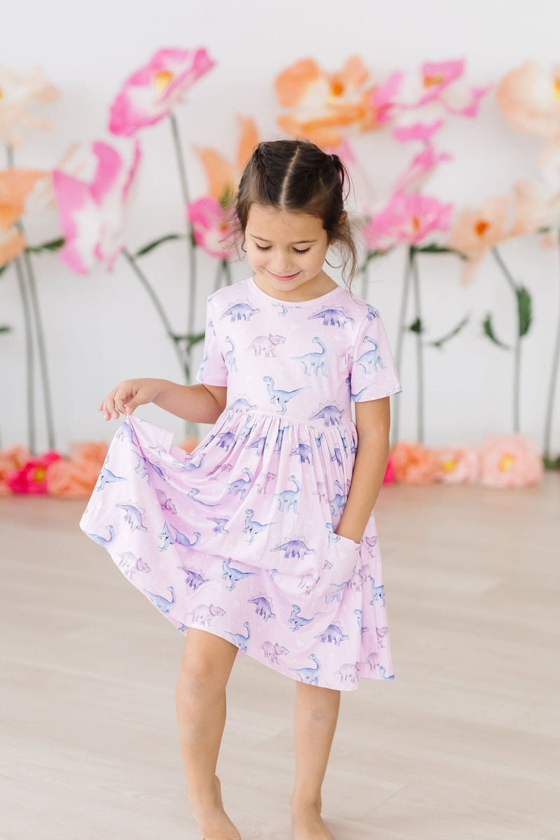 A joyful little girl in the Jurassic Joy S/S Pocket Twirl Dress by Mila & Rose, twirling playfully with a beautiful floral backdrop.