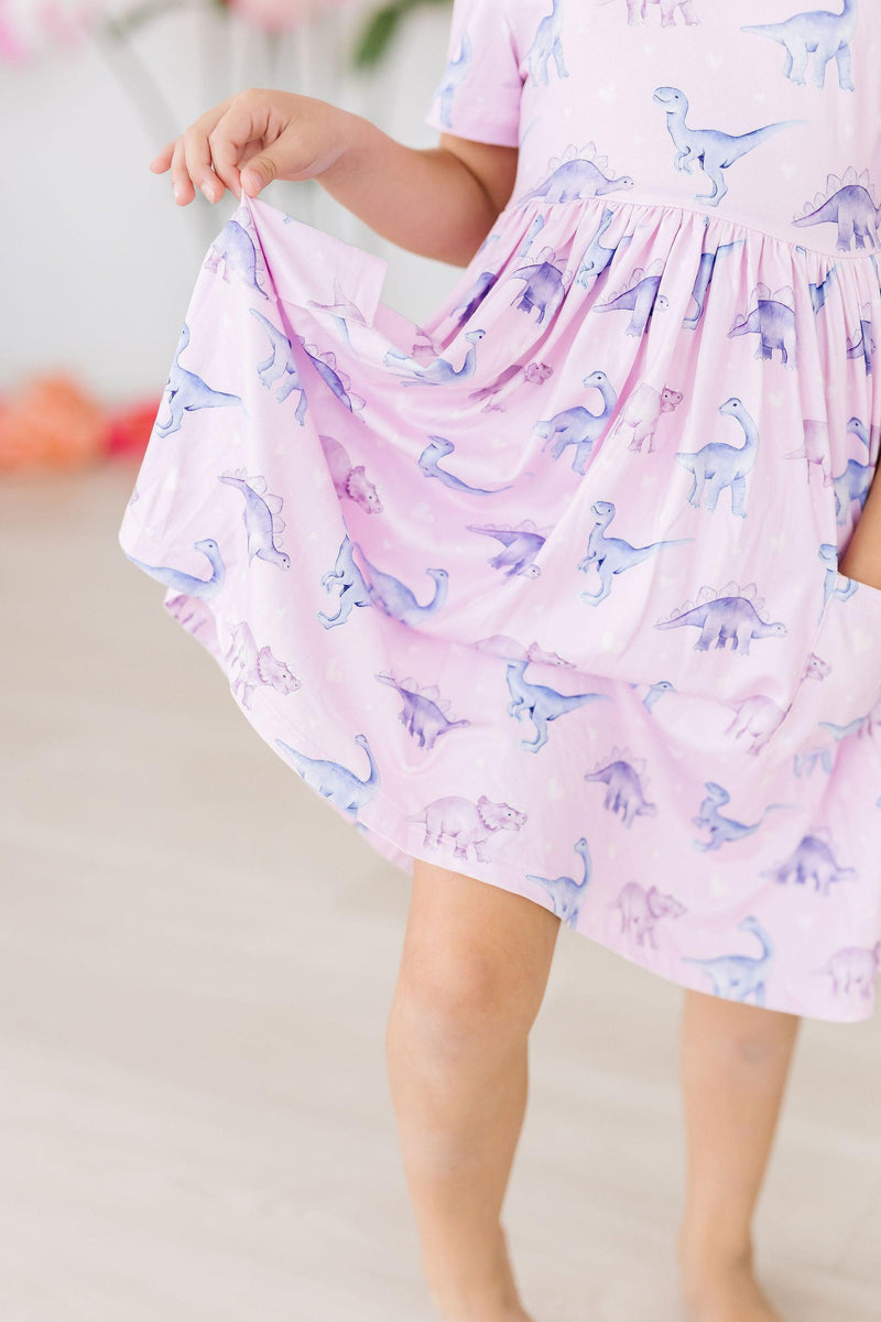 Close-up of a little girl holding the hem of her Jurassic Joy S/S Pocket Twirl Dress by Mila & Rose, featuring a vibrant dinosaur print.