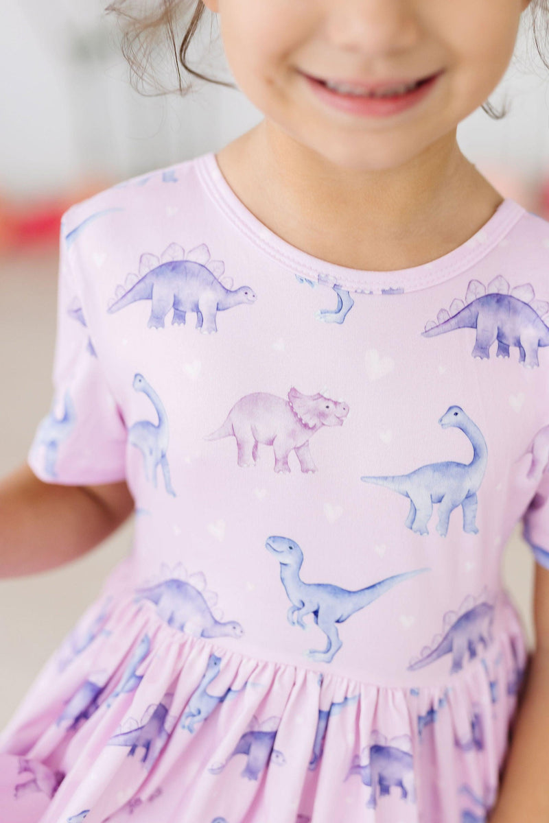 A smiling child in the Jurassic Joy S/S Pocket Twirl Dress by Mila & Rose, highlighting the cute dinosaur patterns and soft fabric.