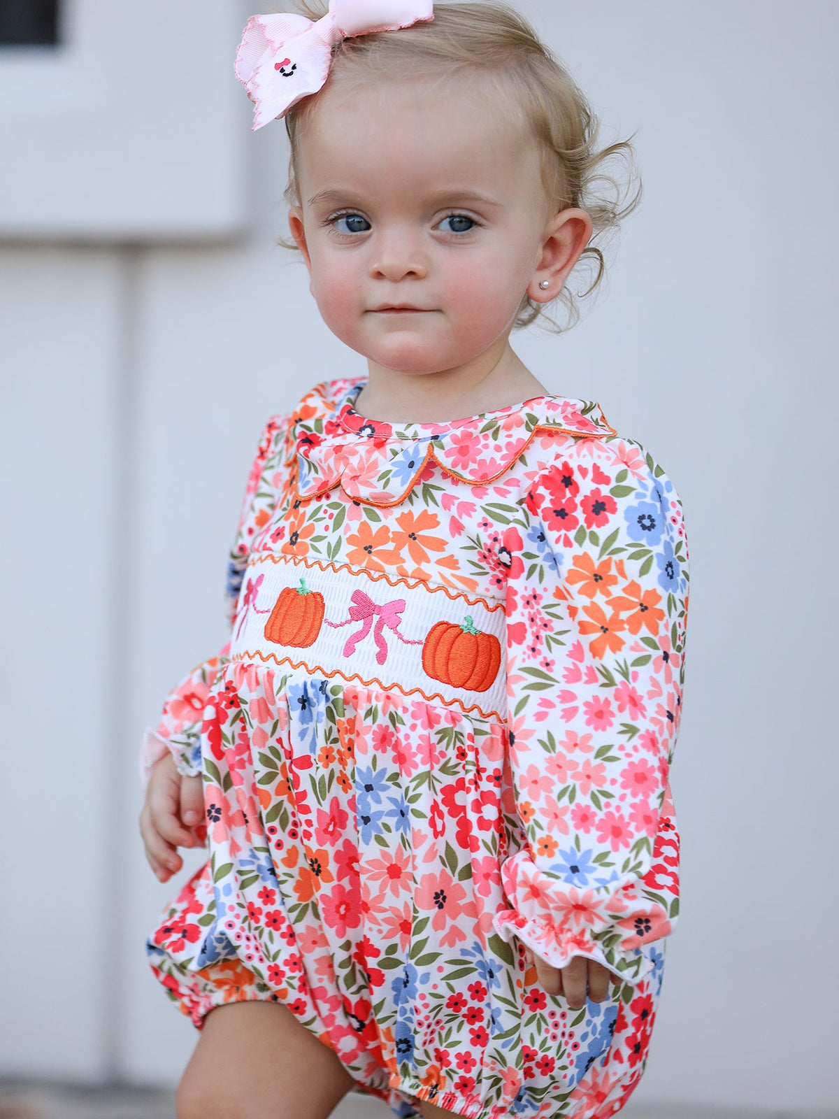Toddler girl wearing a fall floral pumpkin-embroidered smock romper with a pink bow headband.