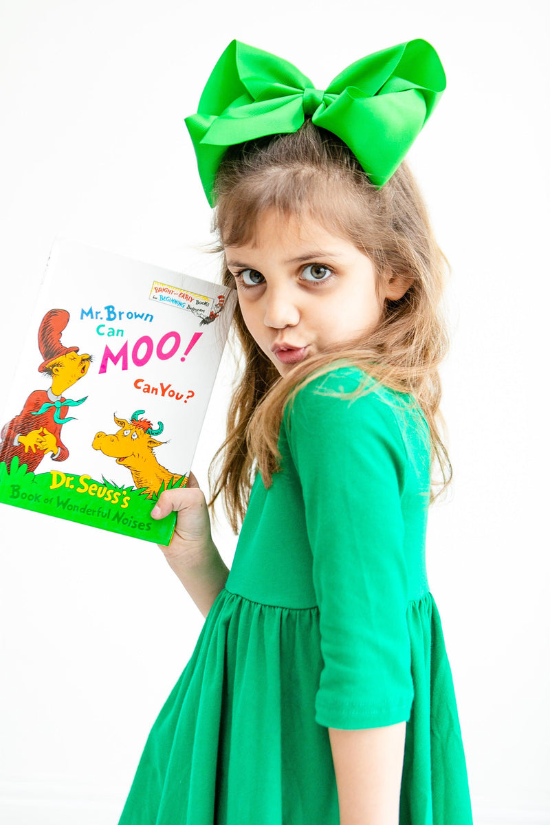 Adorable model wearing the Kelly Green 3/4 Sleeve Pocket Twirl Dress, posing playfully with a Dr. Seuss book, accentuated by a matching green bow, embodying fun and style.