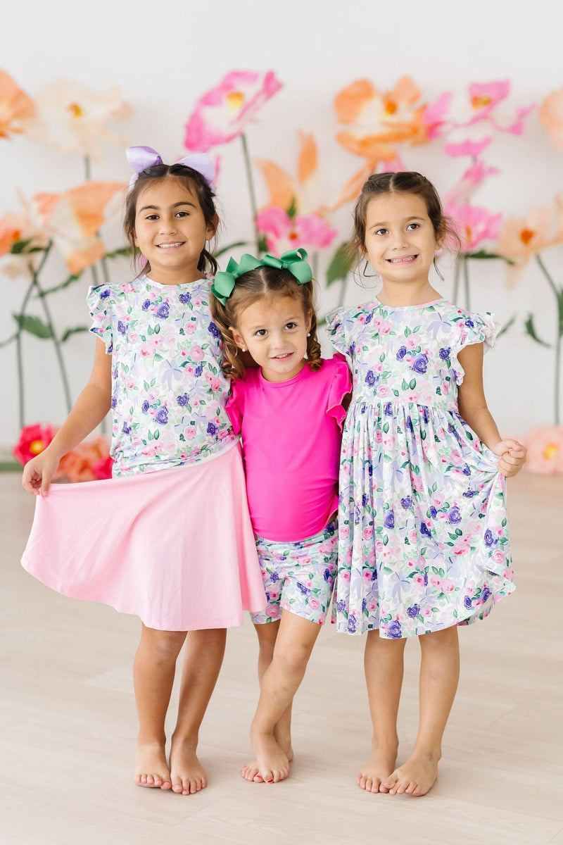 Three cheerful little girls wearing the Lavender Love S/S Ruffle Tee and other matching outfits, perfect for sisterly twirling and fabulous memories.