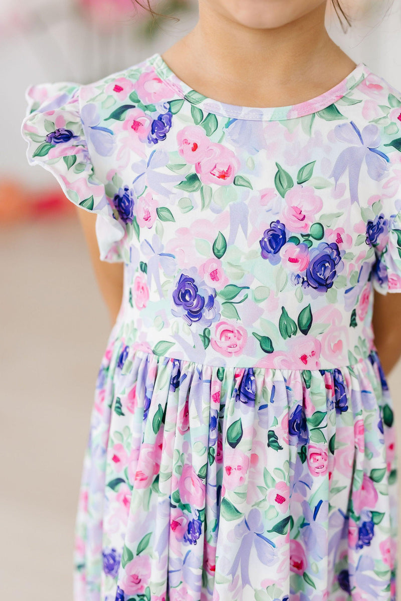 A joyful child wearing the Lavender Love Flutter Sleeve Twirl Dress, showcasing its beautiful floral print adorned with lovely pink and purple hues, perfect for playful twirls and everyday adventures.