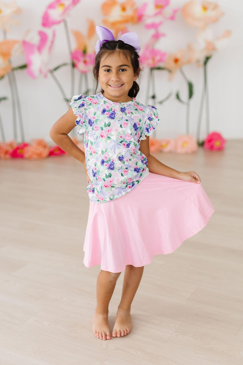 Smiling girl in the Lavender Love S/S Ruffle Tee, radiating joy as she twirls in a pink skirt, embodying a blend of style and fun.