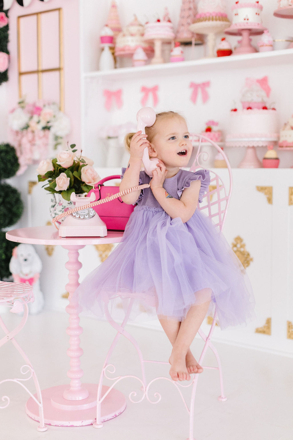 A joyful little girl wearing a Lavender Shimmer Tutu Dress, happily pretending to talk on a vintage phone at a whimsical party setup, embodying childhood magic.