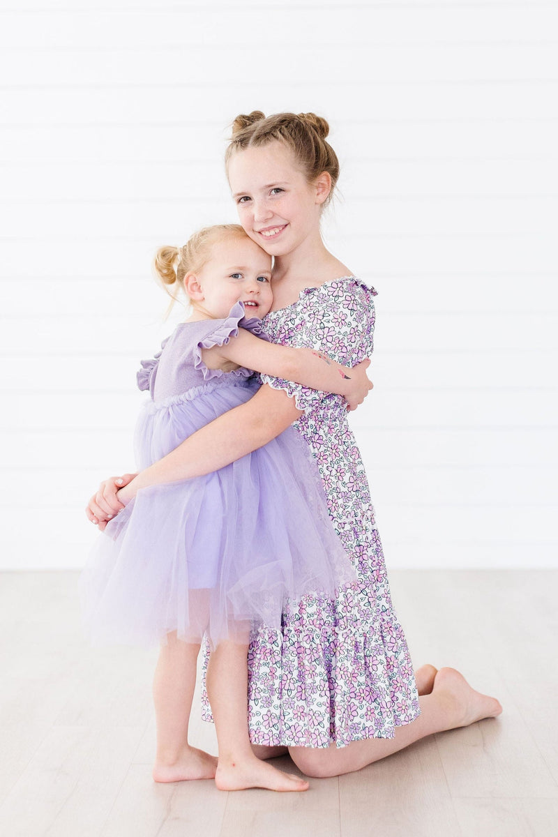 A heartwarming moment of two sisters, the younger in a Lavender Shimmer Tutu Dress, sharing a hug and smiles, revealing the dress's enchanting charm in a minimalistic setting.