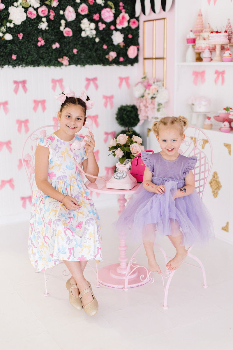 Two adorable girls enjoying a whimsical tea party while wearing the Lavender Shimmer Tutu Dress, radiating joy and charm in a playful setting.