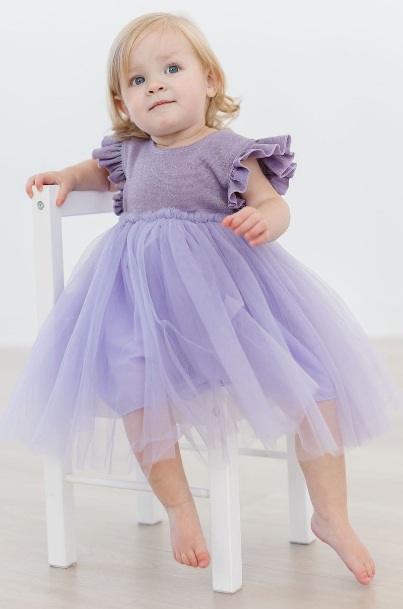 A cute toddler perched on a chair, showcasing the Lavender Shimmer Tutu Dress with flutter sleeves, embodying elegance and playfulness in soft lavender hues.