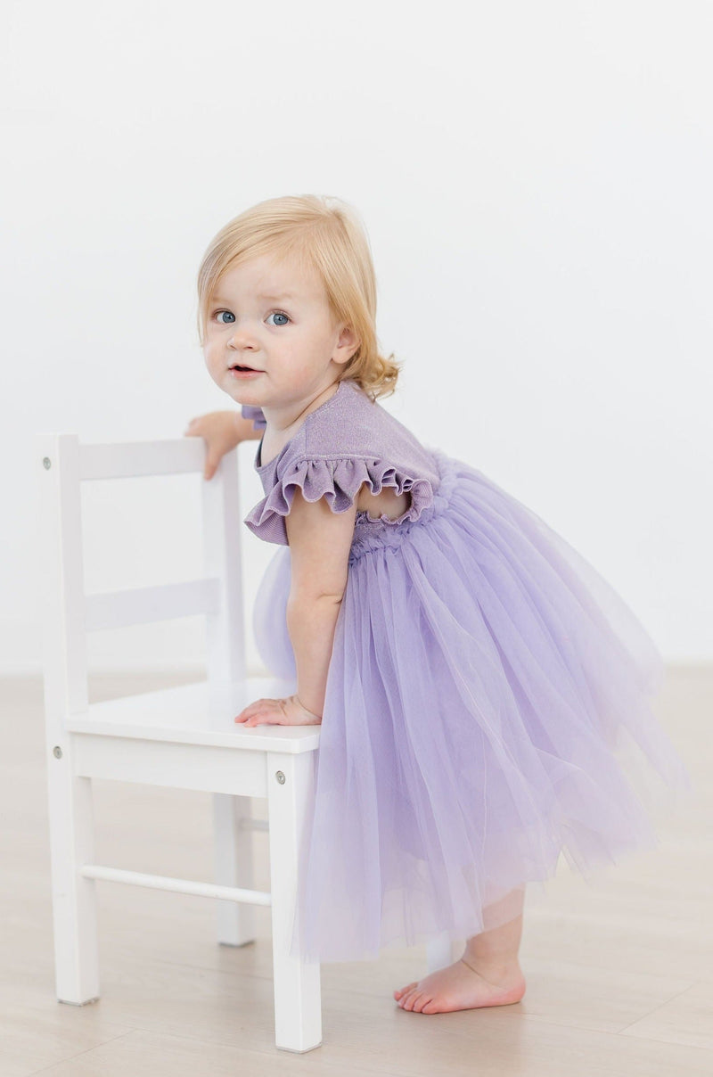 A charming little girl in the Lavender Shimmer Tutu Dress, leaning on a chair and capturing the essence of sweet innocence and sparkling style.