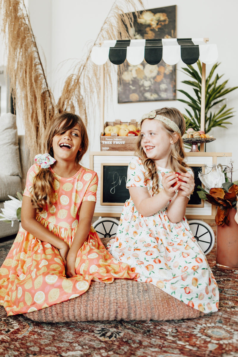 Two children joyfully sporting Lemon Squeezy S/S Pocket Twirl Dresses by Mila & Rose, one in a vibrant lemon pattern and the other in a colorful fruit print, enjoying a fun day.