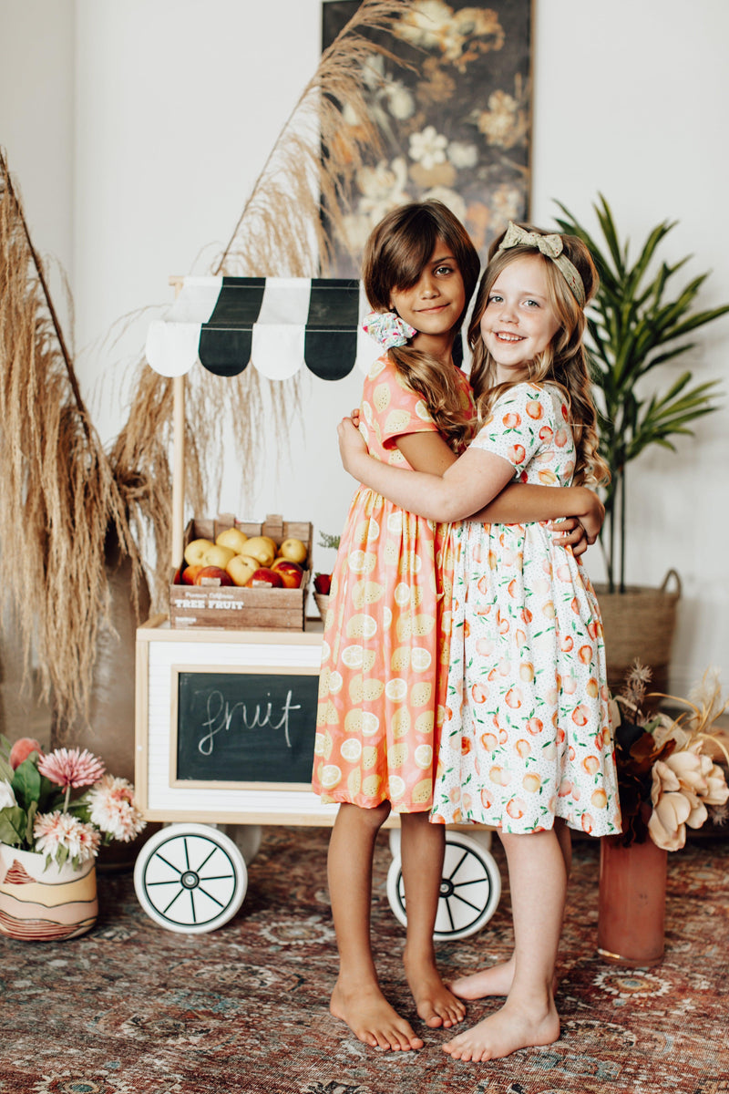 Friends embracing in their Lemon Squeezy S/S Pocket Twirl Dresses by Mila & Rose, showcasing vibrant patterns in a cheerful setting, perfect for playful moments.
