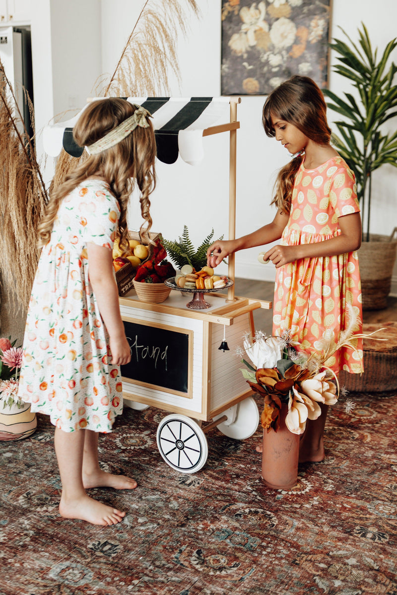 Two girls wearing Lemon Squeezy S/S Pocket Twirl Dress by Mila & Rose® playing at a fruit stand, showcasing the playful designs of the dresses in a lively setting.