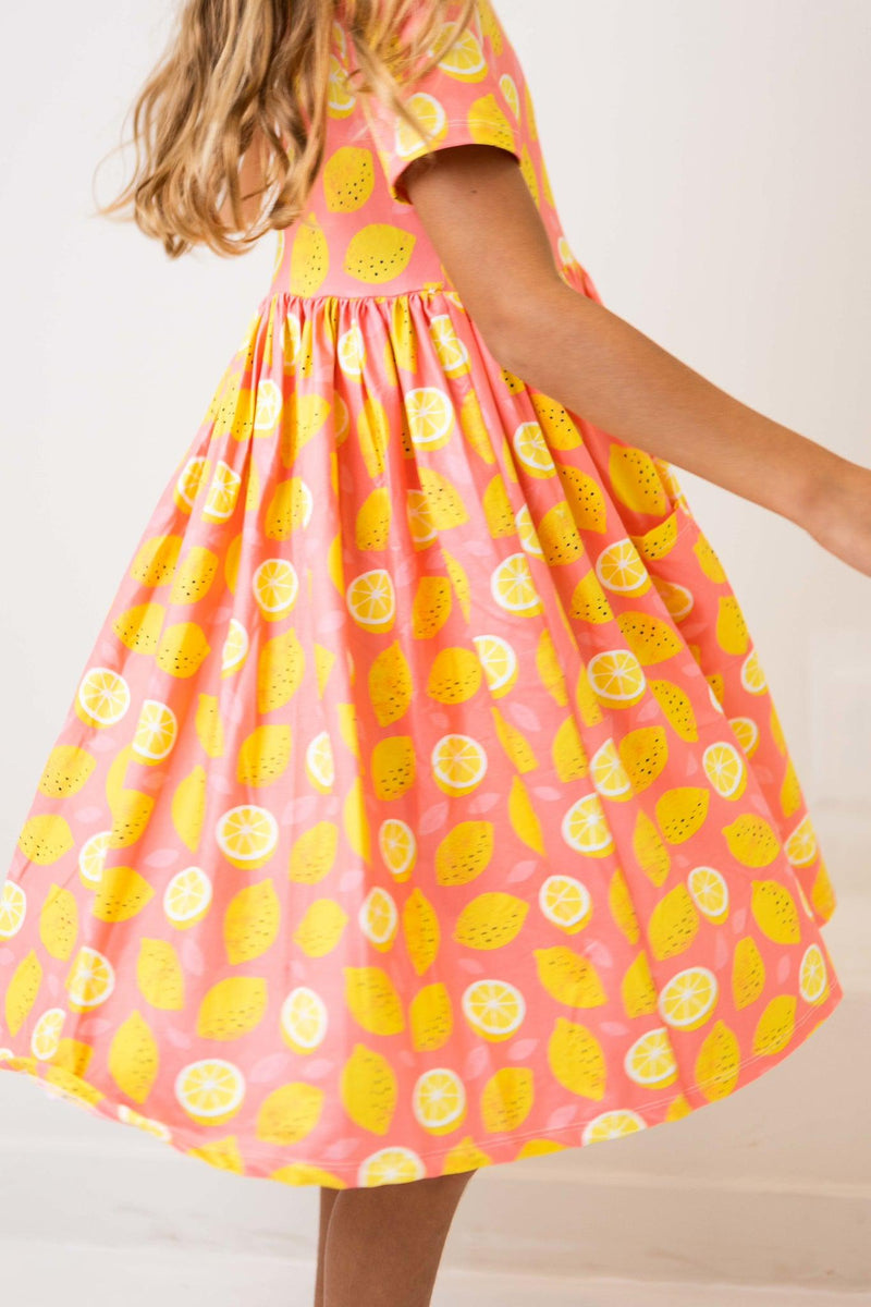 A girl in a Lemon Squeezy S/S Pocket Twirl Dress by Mila & Rose® twirling, highlighting the vibrant lemon pattern of the dress, perfect for play and fun.