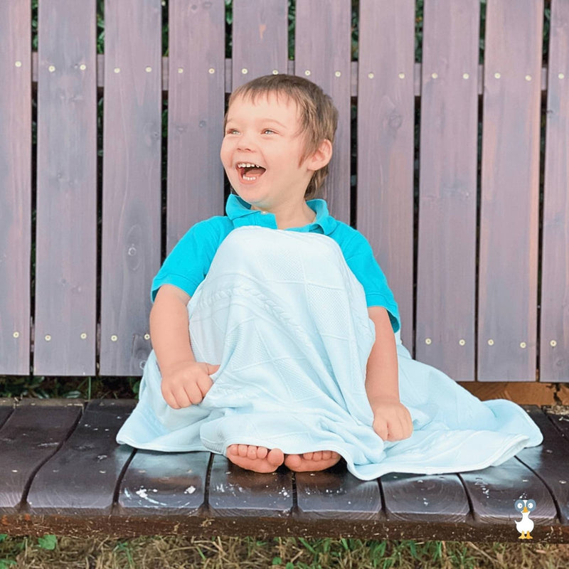 A joyful child sitting on a bench wrapped in the Mint Knit Blanket from Goosewaddle®, demonstrating its playful versatility.