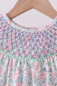 Close-up of a children's dress hanging on a wooden hanger, featuring a smocked multicolor bodice and a white floral skirt with pink, green, and blue flowers.