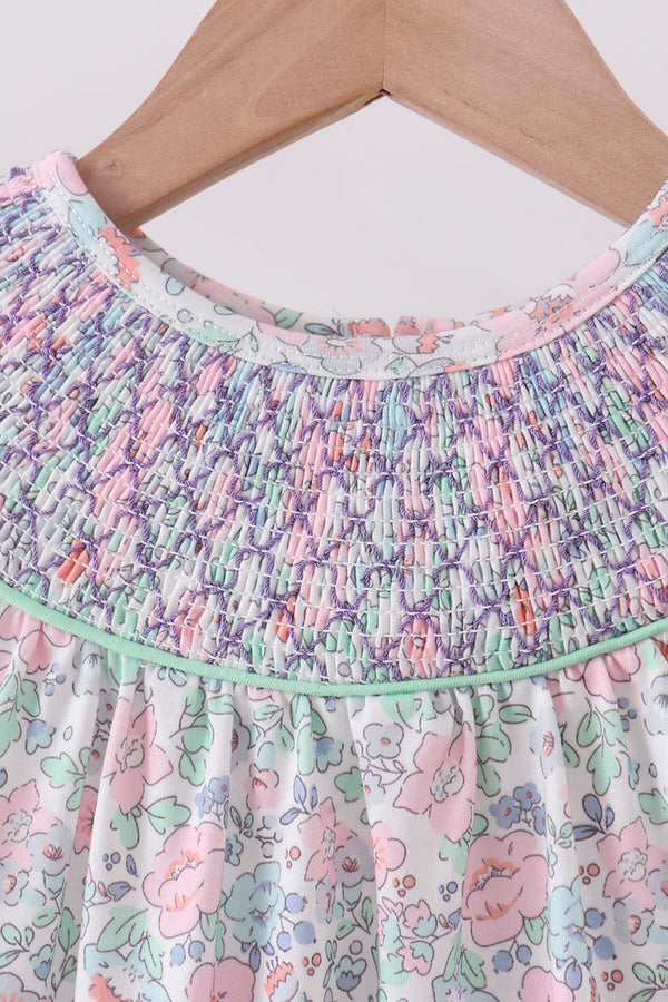 Close-up of a children's dress hanging on a wooden hanger, featuring a smocked multicolor bodice and a white floral skirt with pink, green, and blue flowers.