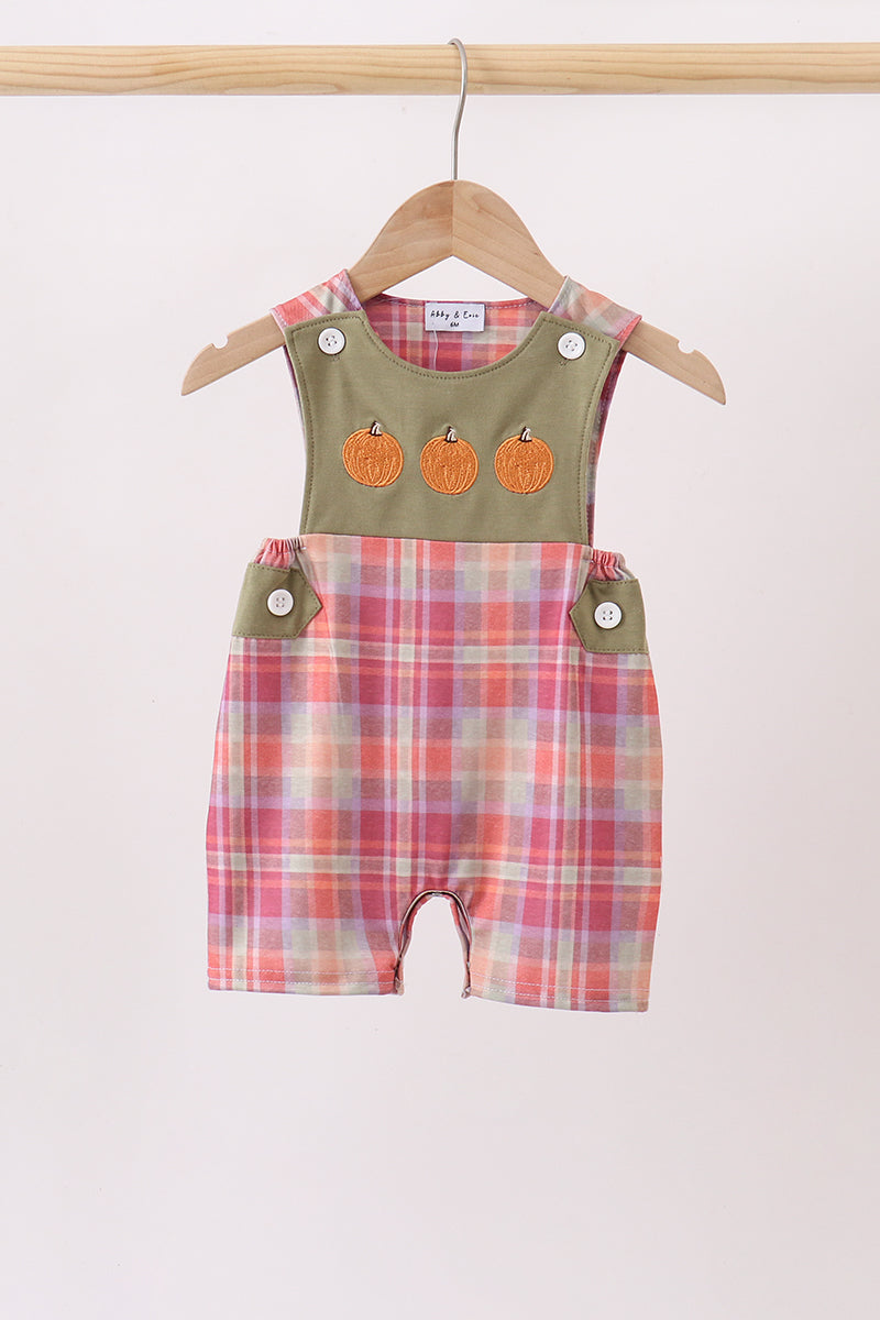 A baby romper with a green solid upper section featuring three orange pumpkin appliques, and pink, orange, and white plaid shorts, hanging on a wooden hanger.