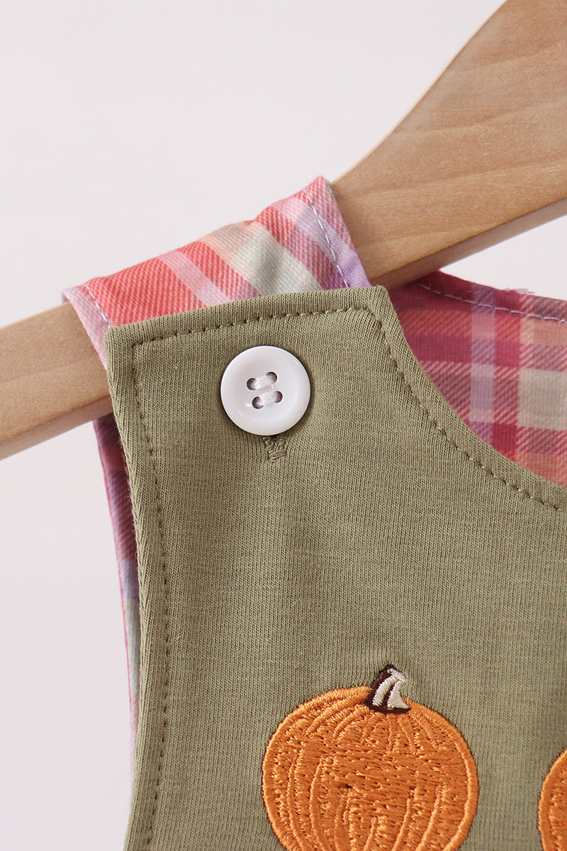 Close-up of a children's sleeveless top or dress in olive green fabric with a white button and embroidered orange pumpkin detail, hanging on a wooden hanger against a light background.