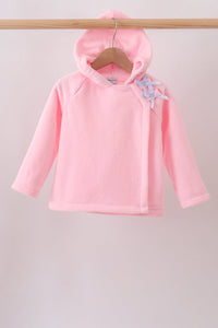 A pink hooded long-sleeve top for a baby or toddler, hanging on a wooden hanger from a wooden clothes rack. The top features a light blue and white patterned bow on the shoulder.