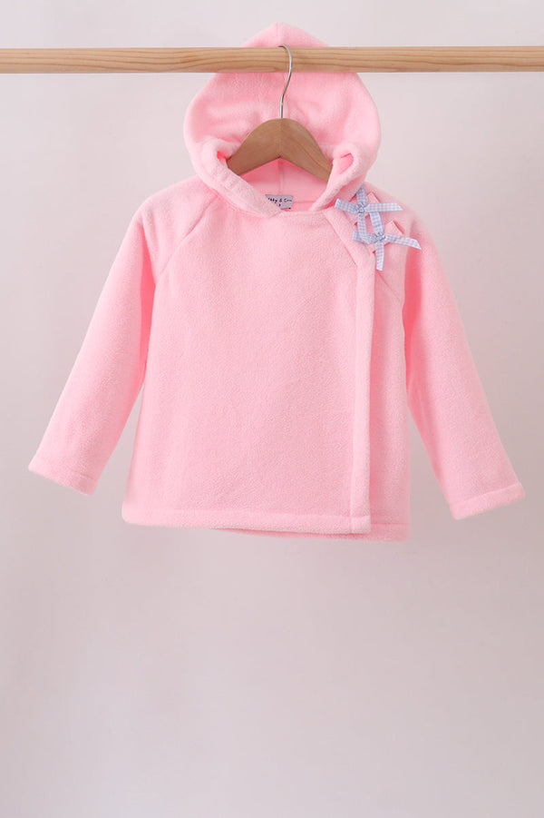 A pink hooded long-sleeve top for a baby or toddler, hanging on a wooden hanger from a wooden clothes rack. The top features a light blue and white patterned bow on the shoulder.
