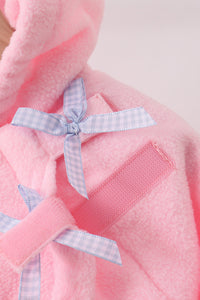 A close-up of a pink plush hooded garment, likely a baby robe or hat, featuring a light blue and white gingham ribbon bow and a pink hook-and-loop strap closure.