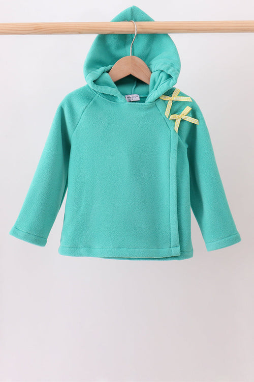 A turquoise long-sleeve hooded sweatshirt hanging on a wooden hanger from a wooden clothes rack, featuring yellow ribbon accents on the shoulder.