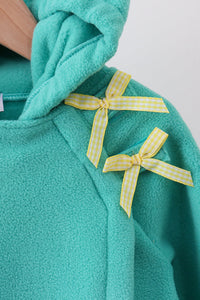 Close-up of a teal fleece hoodie with two yellow-and-white gingham ribbon bows tied on the chest, shown against a light neutral background.