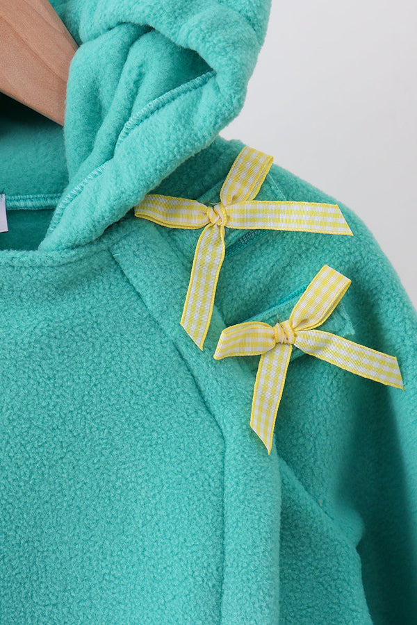 Close-up of a teal fleece hoodie with two yellow-and-white gingham ribbon bows tied on the chest, shown against a light neutral background.
