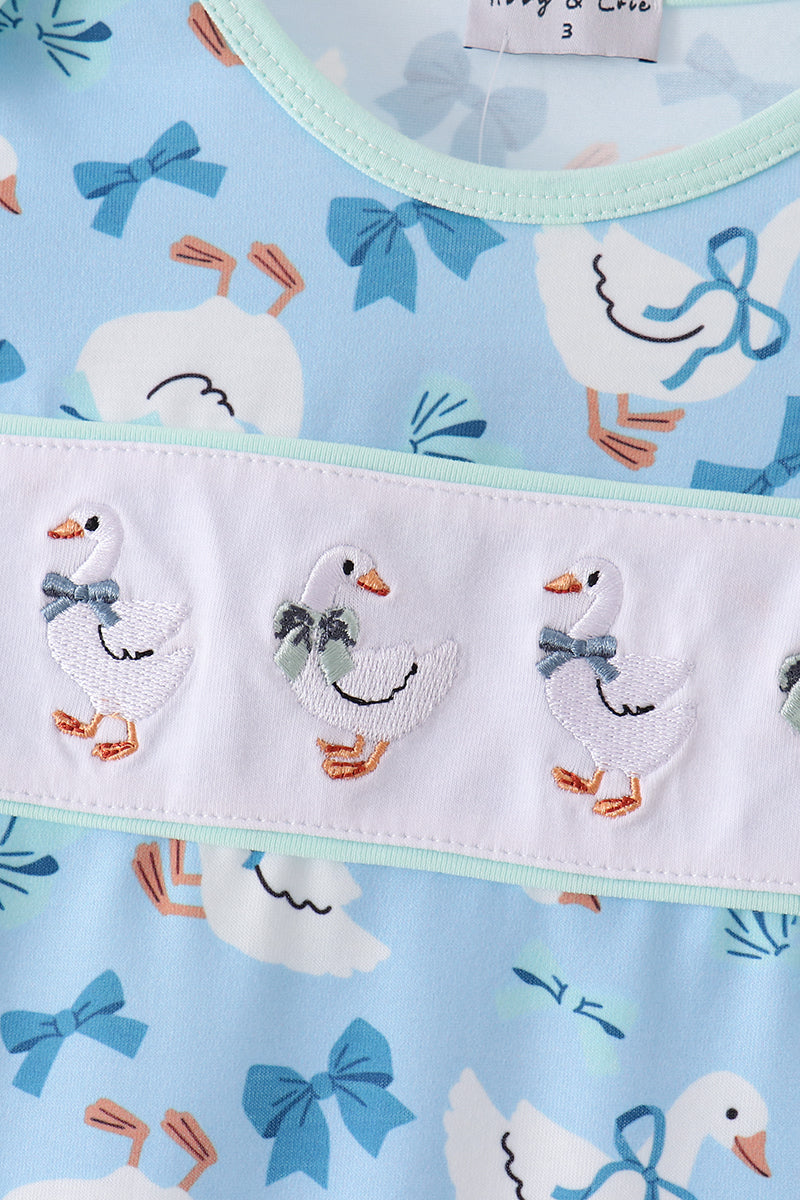 Blue dress featuring embroidered geese with bows on a white ruffle band against a patterned light blue background.