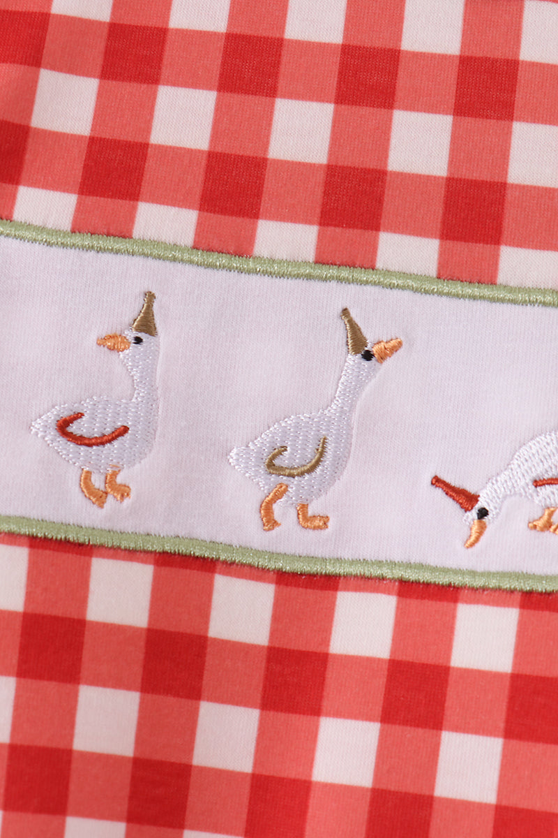 Red gingham fabric featuring embroidered white geese with party hats on a white band with green trim.