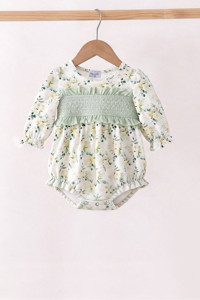 A baby girl's long-sleeve romper with a white and yellow floral print, featuring a smocked light green chest panel and ruffled trim, hanging on a wooden hanger.