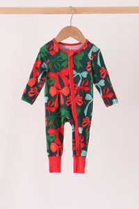 A baby long-sleeve footless one-piece jumpsuit with a red and green holiday bow and leaf pattern, hanging on a wooden hanger against a light beige background.