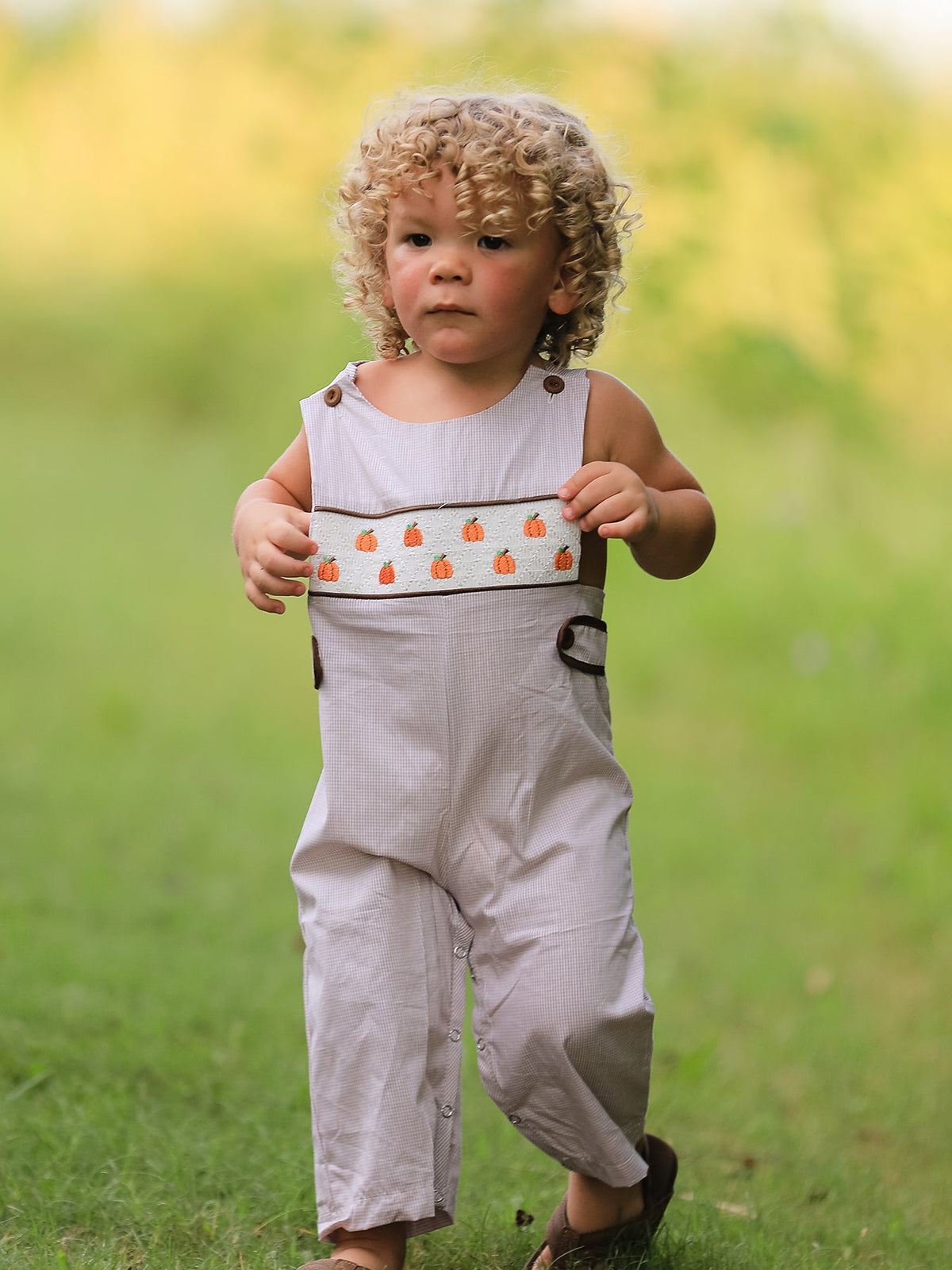 Toddler boy in beige pumpkin-embroidered smocked romper outdoors on a grassy field.
