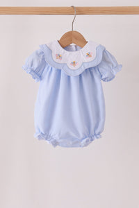A light blue baby romper with a white ruffled collar featuring embroidered pastel flowers, hanging on a wooden hanger against a plain light beige background.