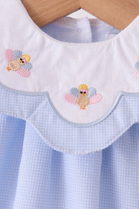 Close-up of a baby dress with a white Peter Pan collar featuring embroidered pastel birds, paired with a light blue gingham skirt, hanging on a wooden hanger.