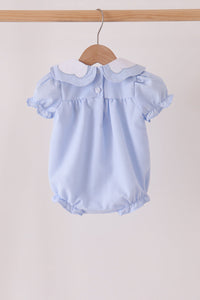 A light blue baby romper with a white Peter Pan collar, hanging on a wooden hanger from a wooden clothes rack against a plain beige background.