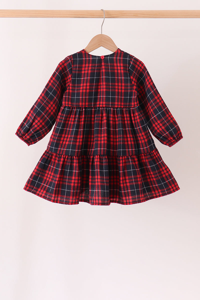 A toddler girl's long-sleeve tiered dress in a red and black plaid pattern, hanging on a wooden hanger from a light wood clothing rack against a plain off-white background.